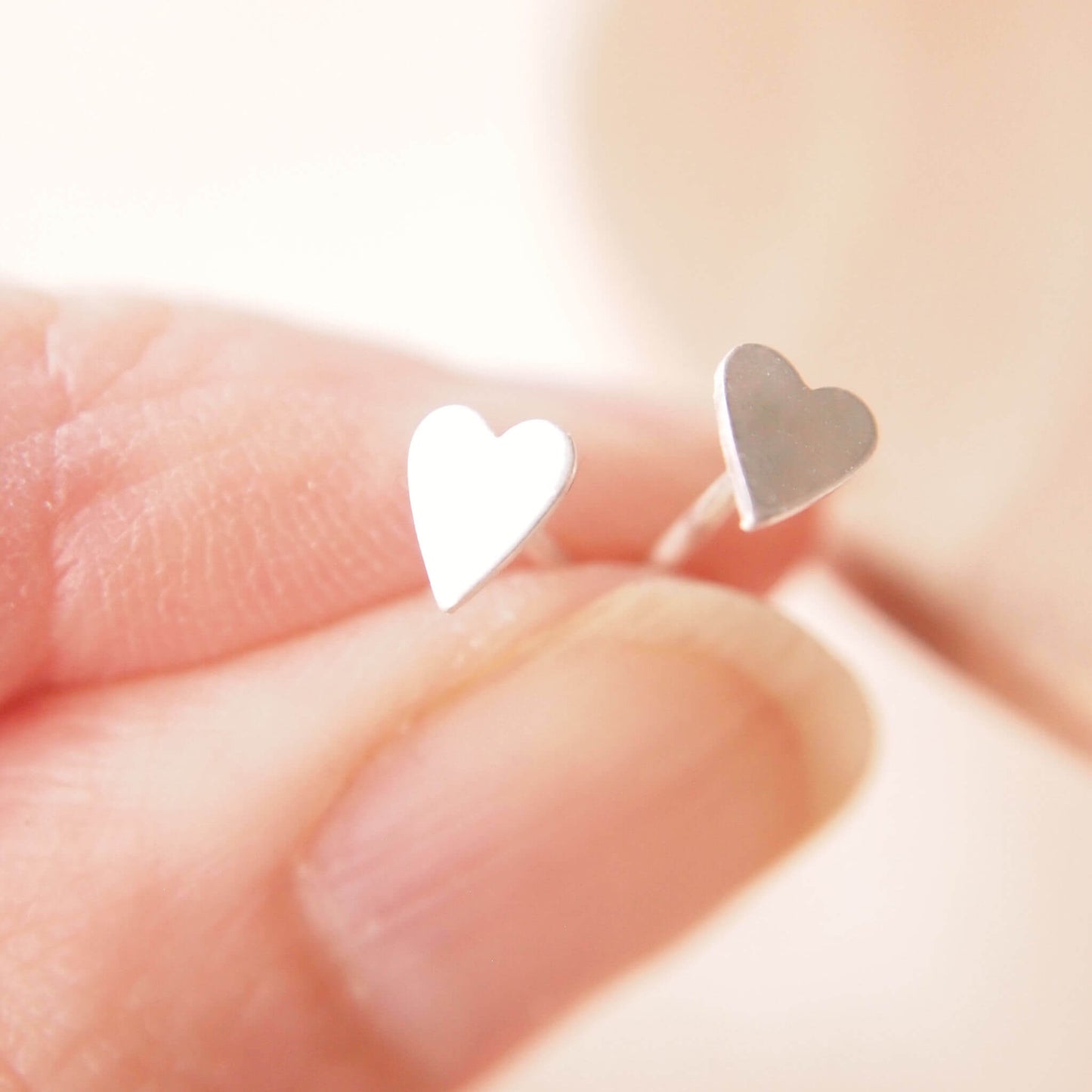 A pair of small heart shaped silver studs made by maram jewellery held between two fingers to show size