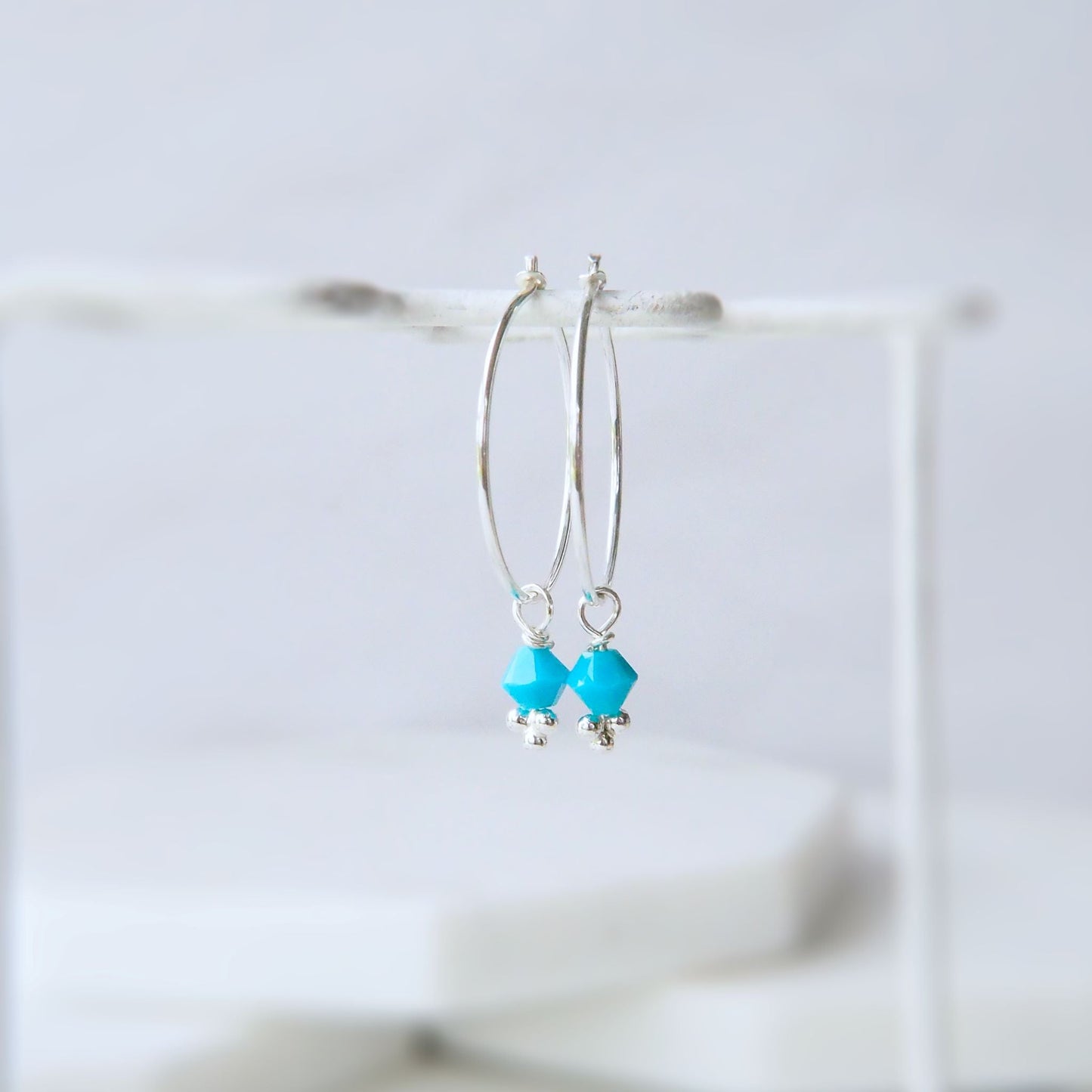 Sterling Silver boho thin hoops with turquoise crystal bicone dropper in December Birthstone. Pictured hanging from a white jewellery rail on a white background. Jewellery Handmade in Edinburgh UK by maram jewellery