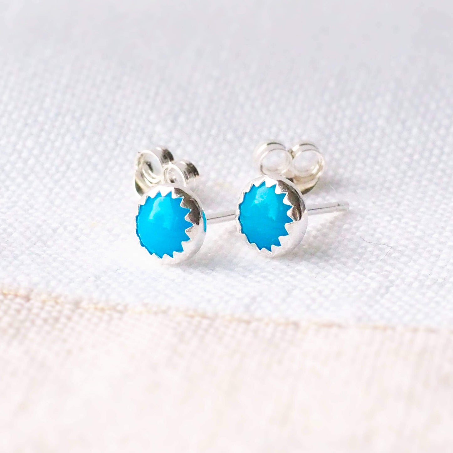 Beautifully simple modern gemstone stud earrings with a turquoise gemstone set by hand into a modern minimalist sterling silver surround, sitting with earring backs on a linen cloth.The earrings are 5mm in size, round and come with butterfly backs.Turquoise is the birthstone for December. These stud earrings are handcrafted by a small business in Scotland UK by designer jeweller Maram Jewellery