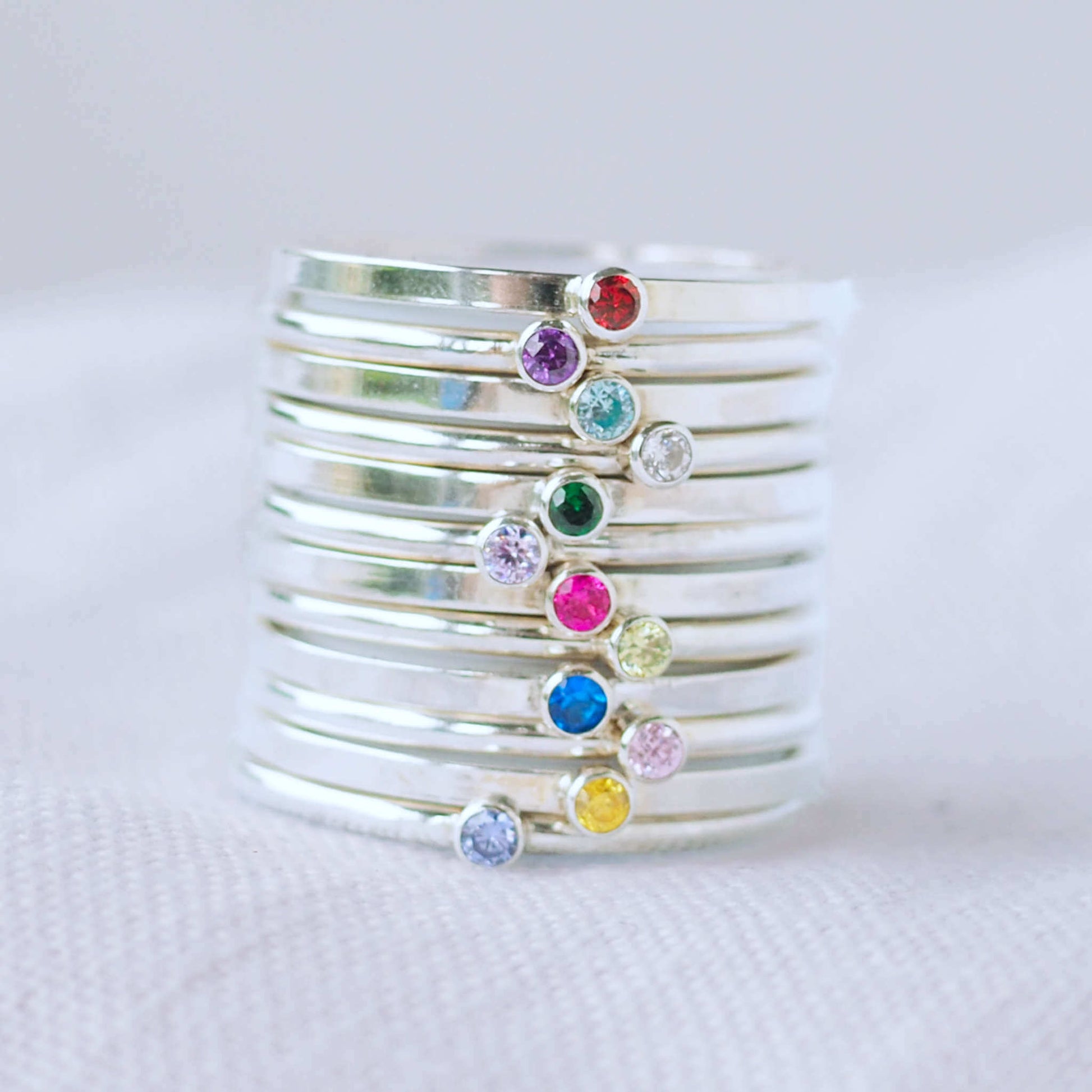 Stack of twelve birthstone rings in Sterling Silver with a round coloured cubic zirconia in 2 mm size to mark birthstones for every month. Rainbow bright coloured gems with round or square silver bands to create a modern minimalist gemstone ring. Handmade in Scotland by maram jewellery