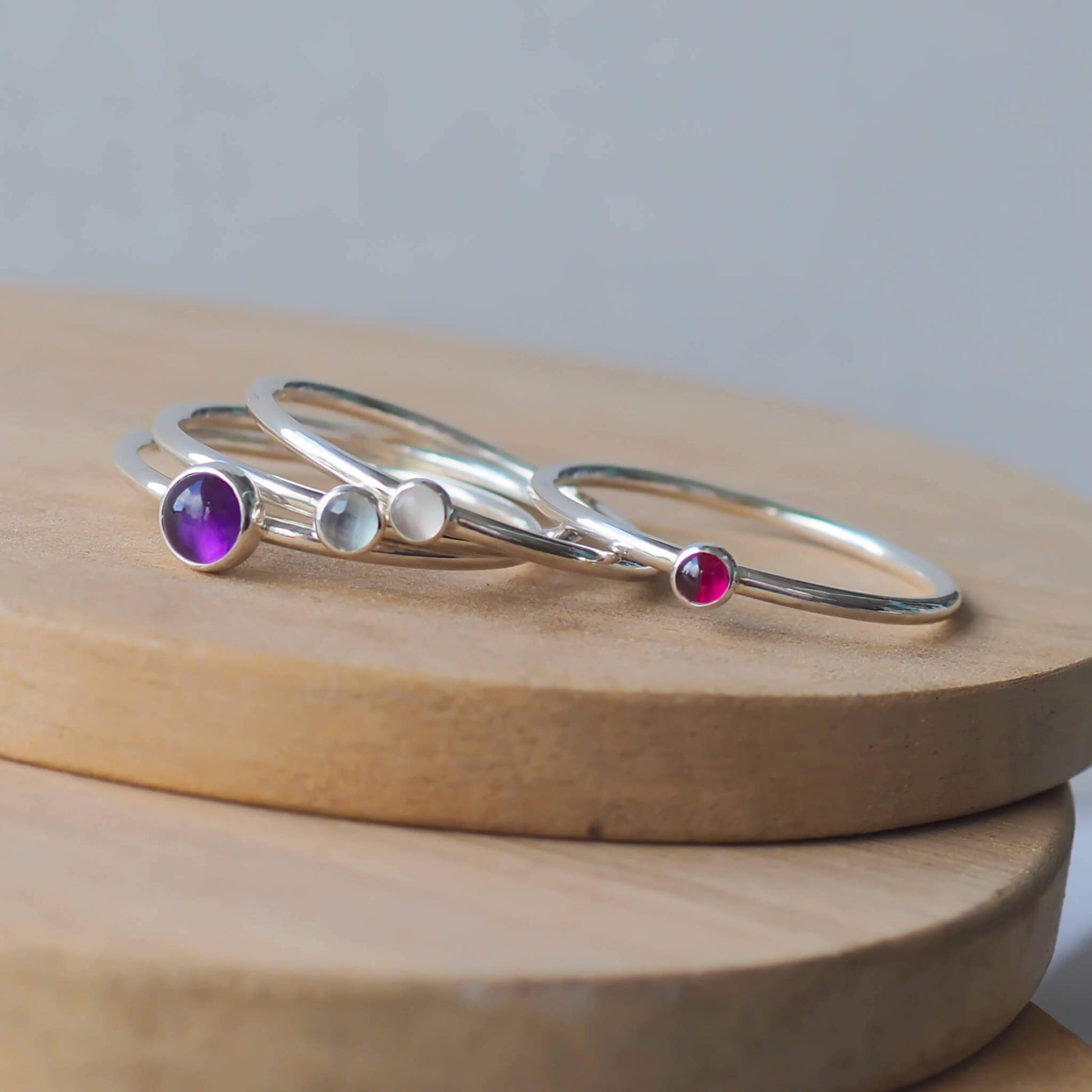 Pile of rings in Sterling Silver and mixed gemstones. The rings are minimalist in style with no decoration and feature a single simple round cabochon in a large 5mm Purple Amethyst, smaller 3mm Blue Topaz, White Moonstone, and rich red Garnet. Handmade in Scotland by maram jewellery