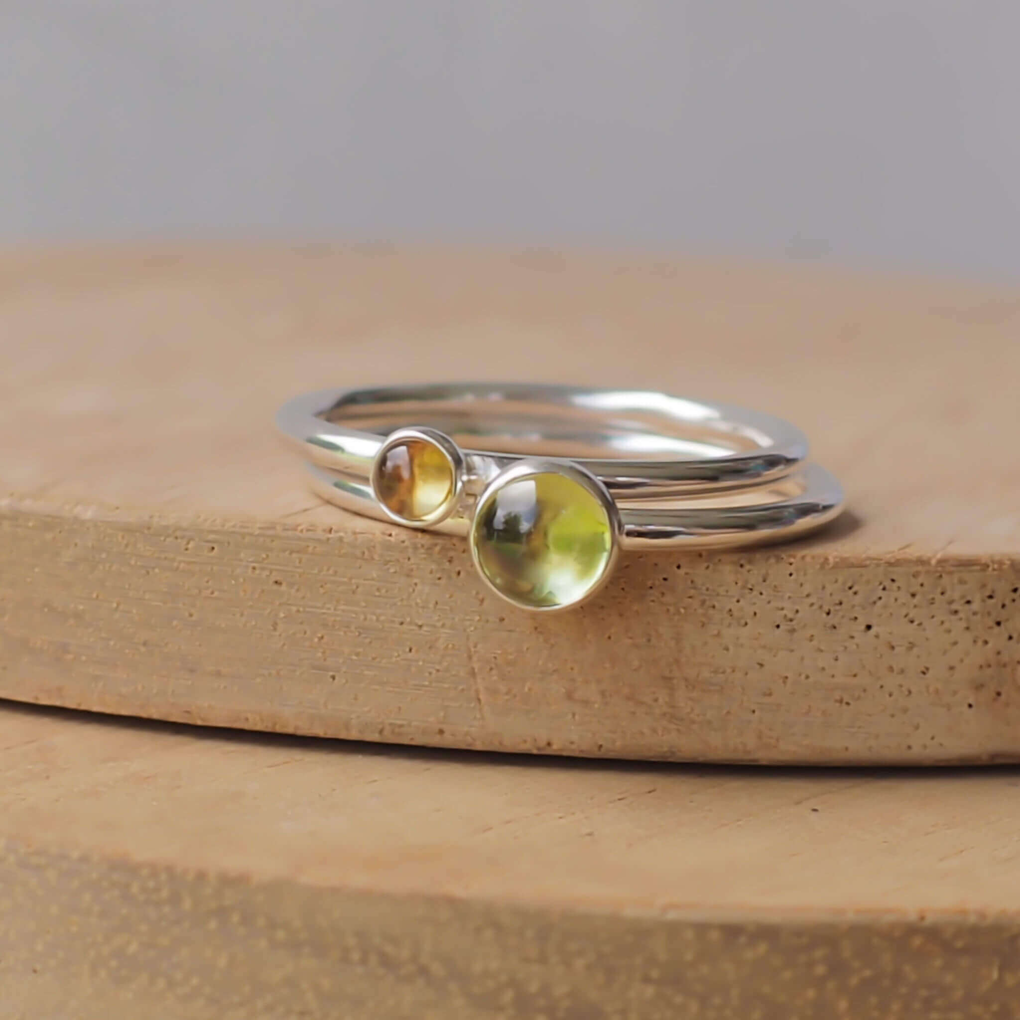 Two silver ring set with round stones. Simple round bands with a 5mm green Peridot and a 3mm yellow Citrine, birthstones for August and November. Rings are pictured on wood background. Handmade by Maram Jewellery in Scotland