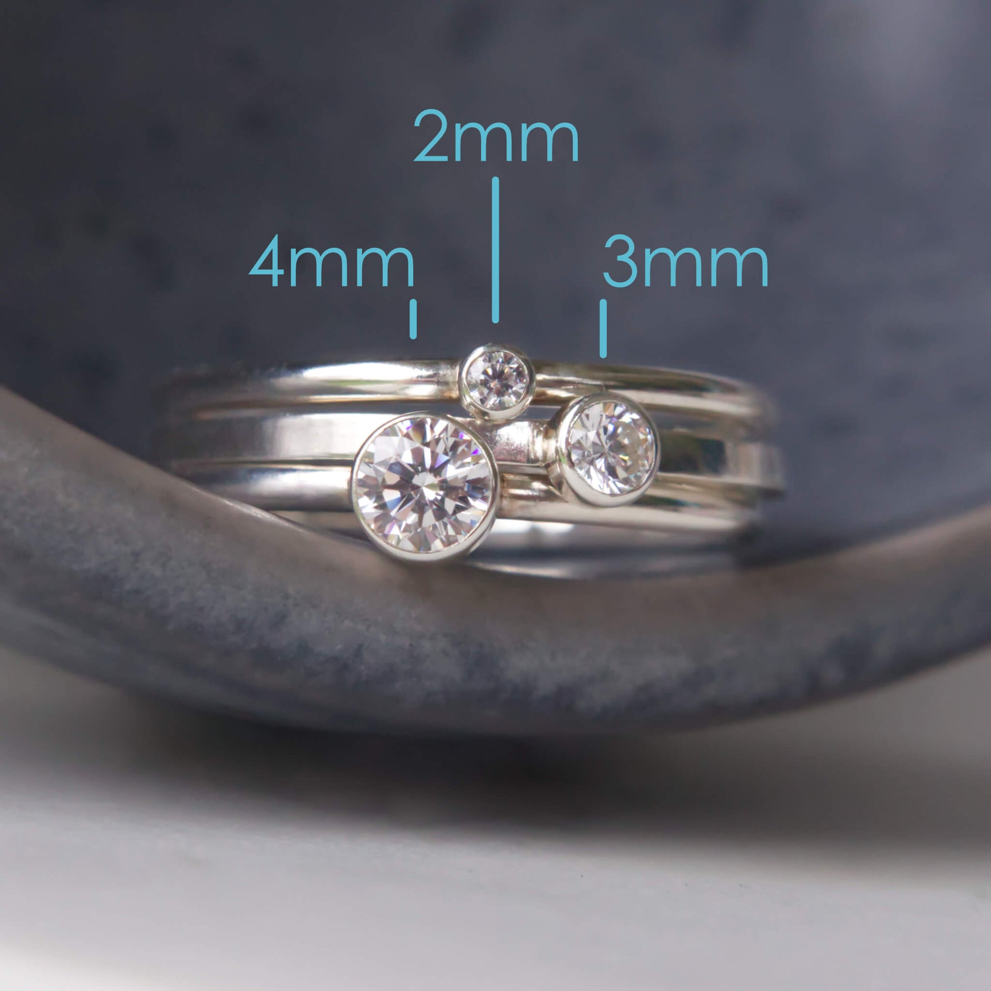 Trio of Diamond Silver Cubic Zirconia Solitaire rings with round stones. Each of the rings have a different sized gem in a 2mm, 3mm and 4mm size, with text to indicate size. Pictured on a grey ceramic background. RIngs handmade by maram jewellery in Scotland UK