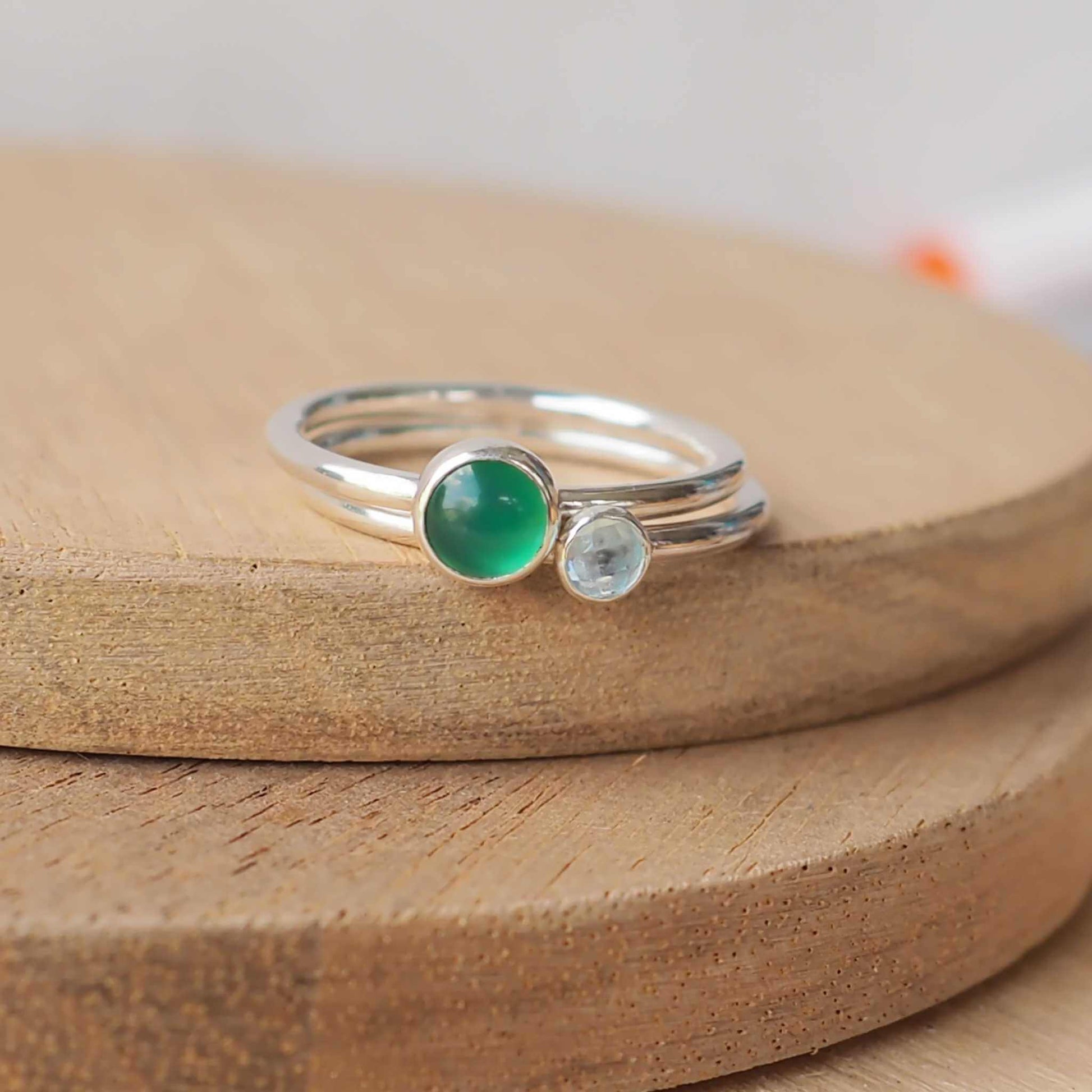 Two silver ring set with round stones. Simple round bands with a 5mm Green Agate and a 3mm Blue Topaz. Rings are pictured on wood background. Handmade in Scotland by Maram Jewellery