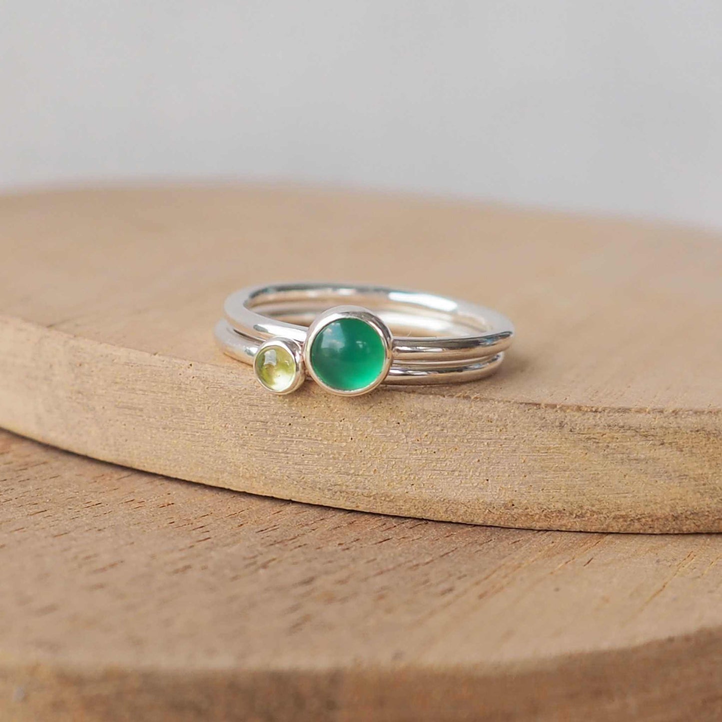 Two silver ring set with round stones. Simple round bands with a 5mm Green Agate and a 3mm Peridot. Rings are pictured on wood background. Handmade in Scotland by Maram Jewellery