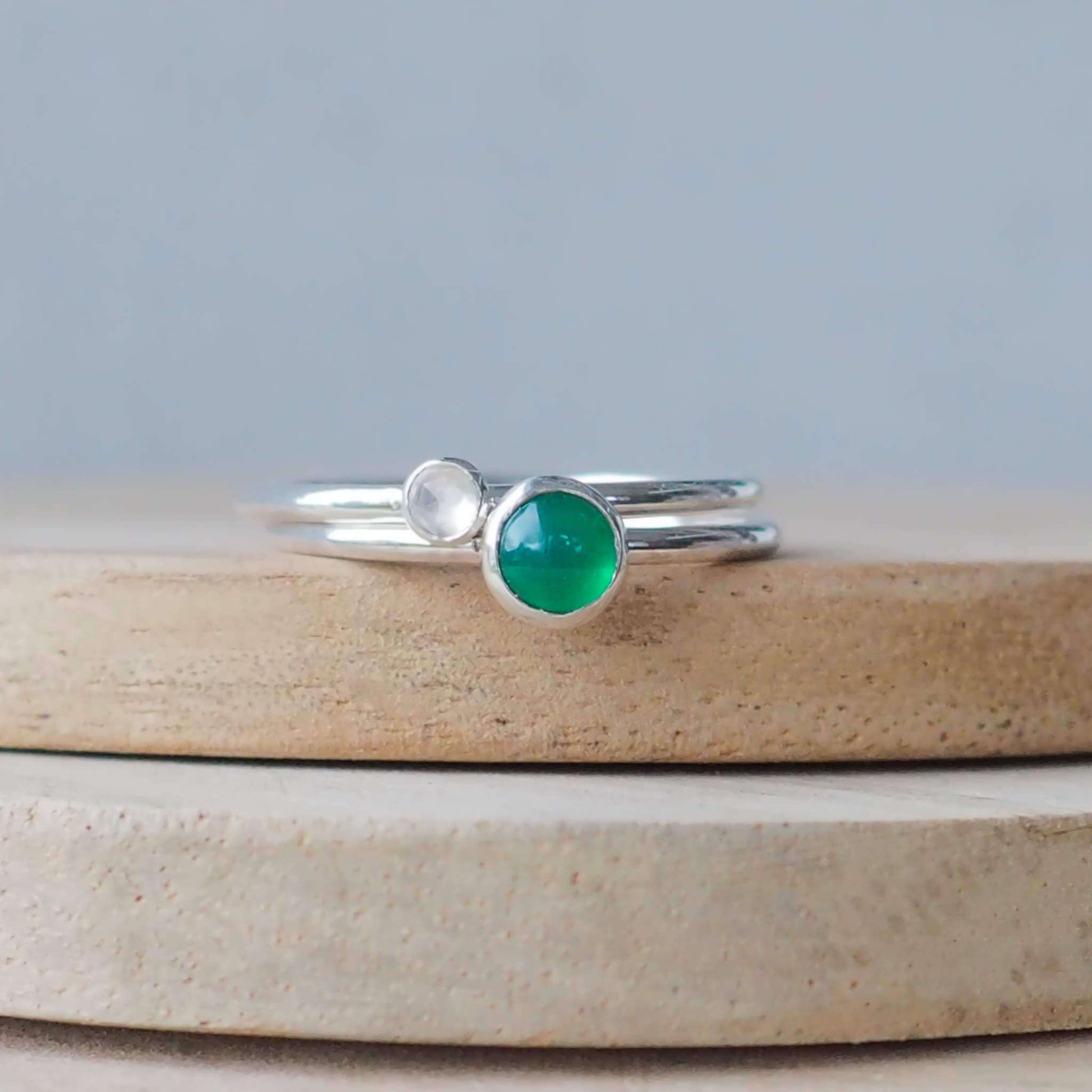 Two silver ring set with round stones. Simple round bands with a 5mm Green Agate and a 3mm White Topaz. Rings are pictured on wood background. Handmade in Scotland by Maram Jewellery