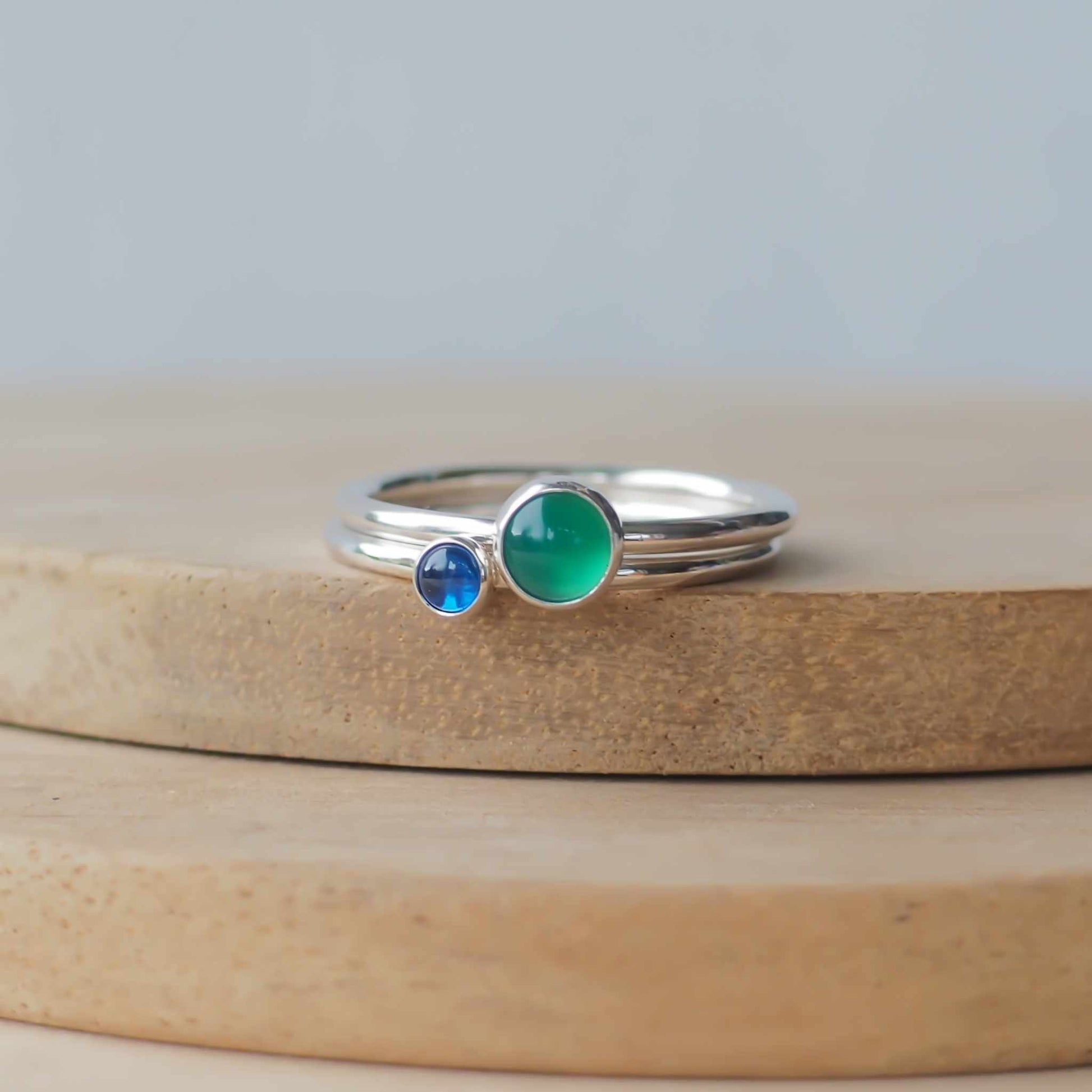 Two silver ring set with round stones. Simple round bands with a 5mm Green Agate and a 3mm Lab Sapphire. Rings are pictured on wood background. Handmade in Scotland by Maram Jewellery