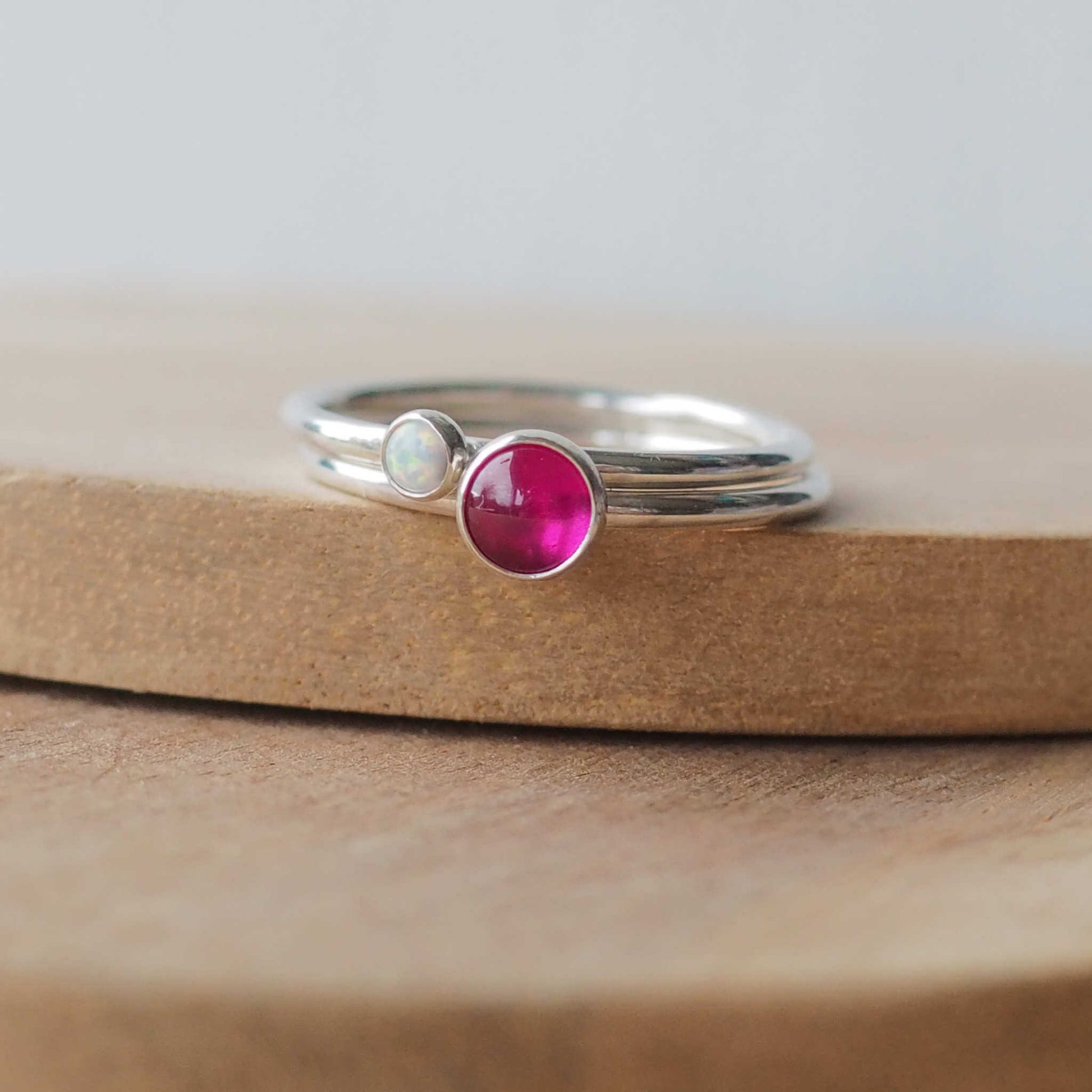 Two silver ring set with round stones. Simple round bands with a 5mm pinky red lab Ruby and a 3mm  Lab Opal. Rings are pictured on wood background. Handmade by Maram Jewellery in Scotland