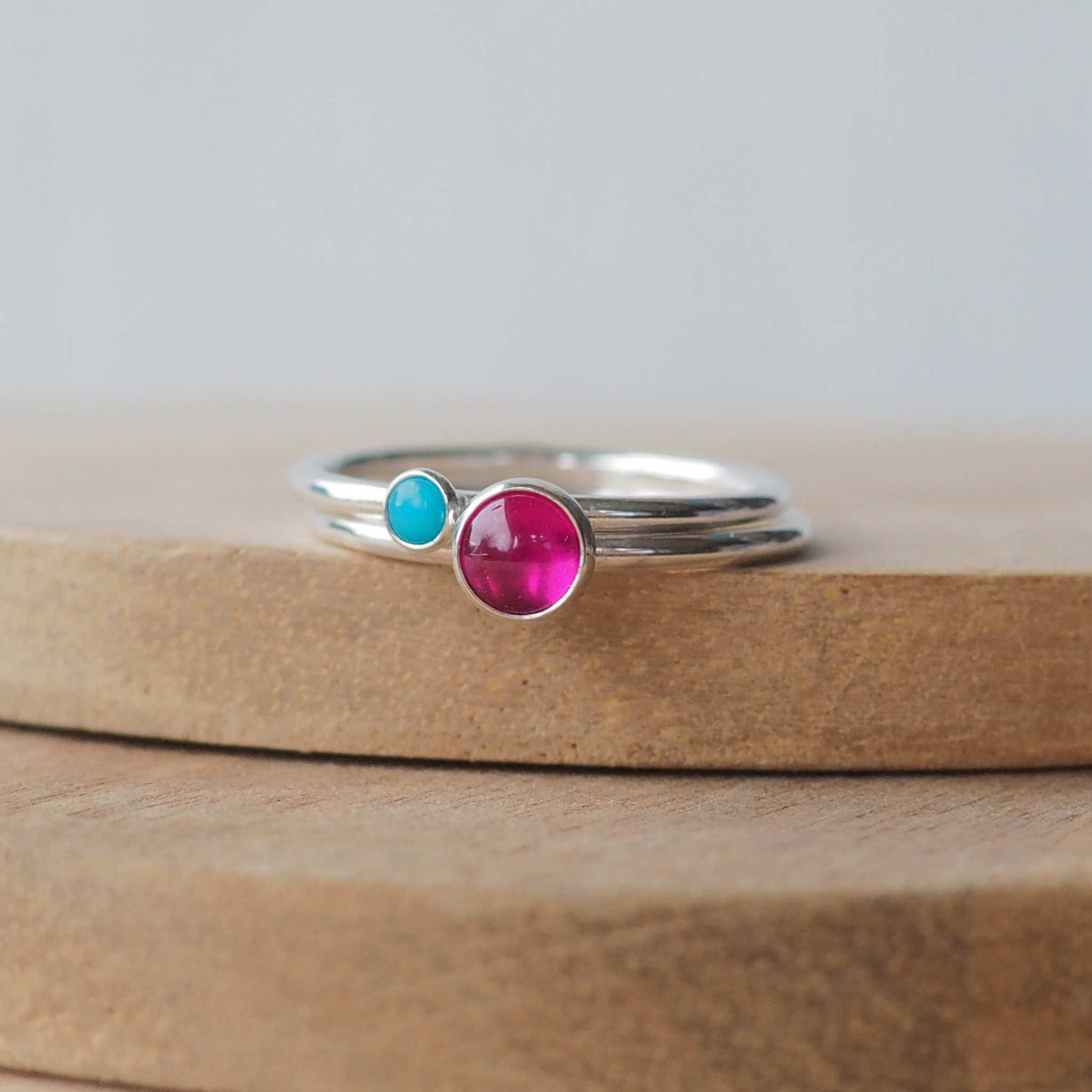 Two silver ring set with round stones. Simple round bands with a 5mm pinky red lab Ruby and a 3mm  Turquoise. Rings are pictured on wood background. Handmade by Maram Jewellery in Scotland
