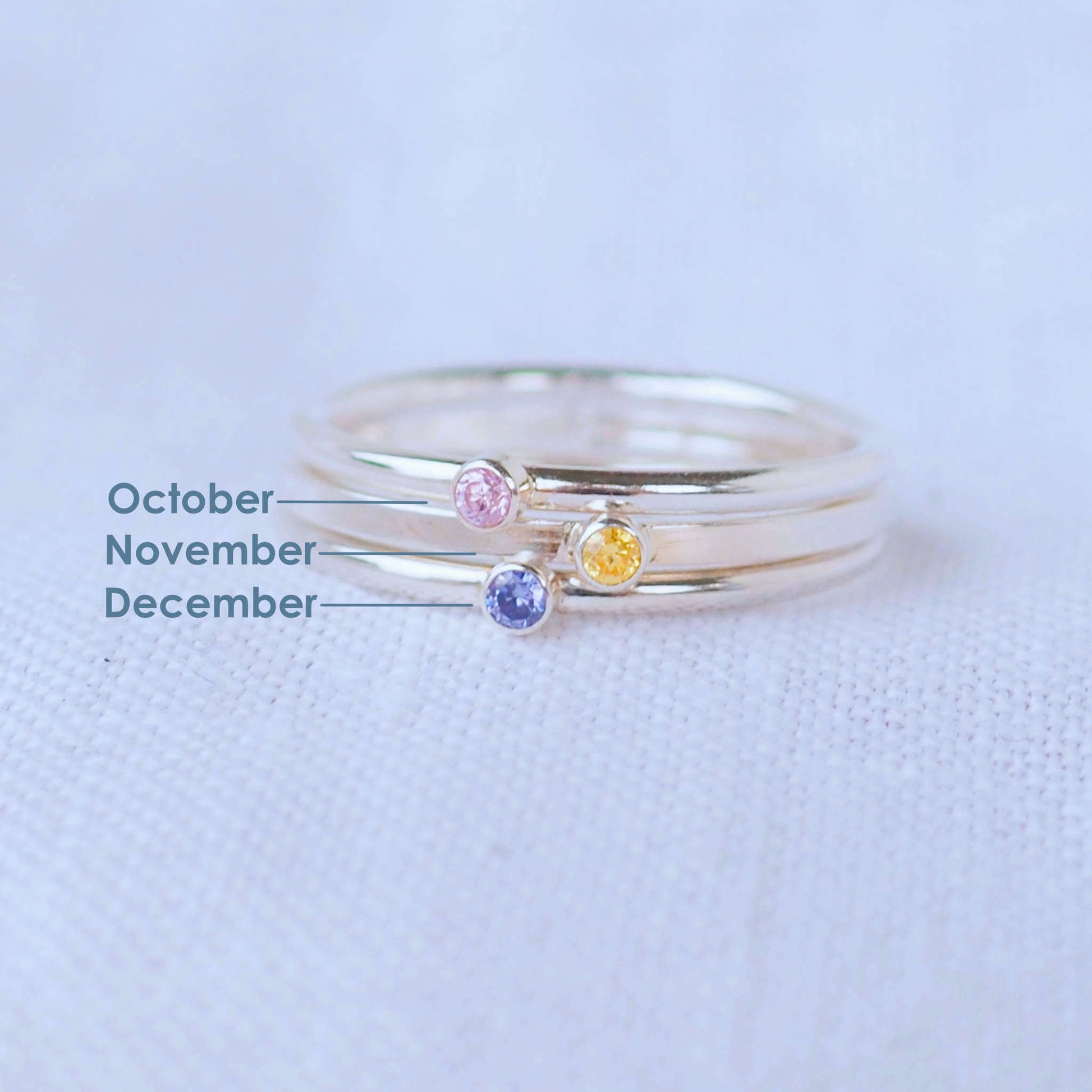 Three silver rings showing October, November and December  Birthstones. The rings are simple silver bands with a miniature birthstone on the band in pale pink, yellow and violet cubic zirconia. Handmade in Edinburgh by maram jewellery