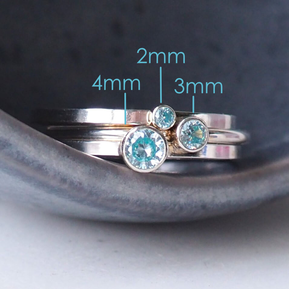 Set of three Aquamarine cubic zirconia rings stacked in a grey ceramic bowl. With text to mark the three sizes - 2mm, 3mm and 4mm round gemstones.Handmade by maram jewellery in Scotland UK