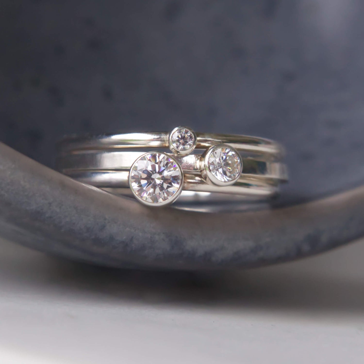 Trio of Diamond Silver Cubic Zirconia Solitaire rings with round stones. Each of the rings have a different sized gem in a 2mm, 3mm and 4mm size, with text to indicate size. Pictured on a grey ceramic background. RIngs handmade by maram jewellery in Scotland UK