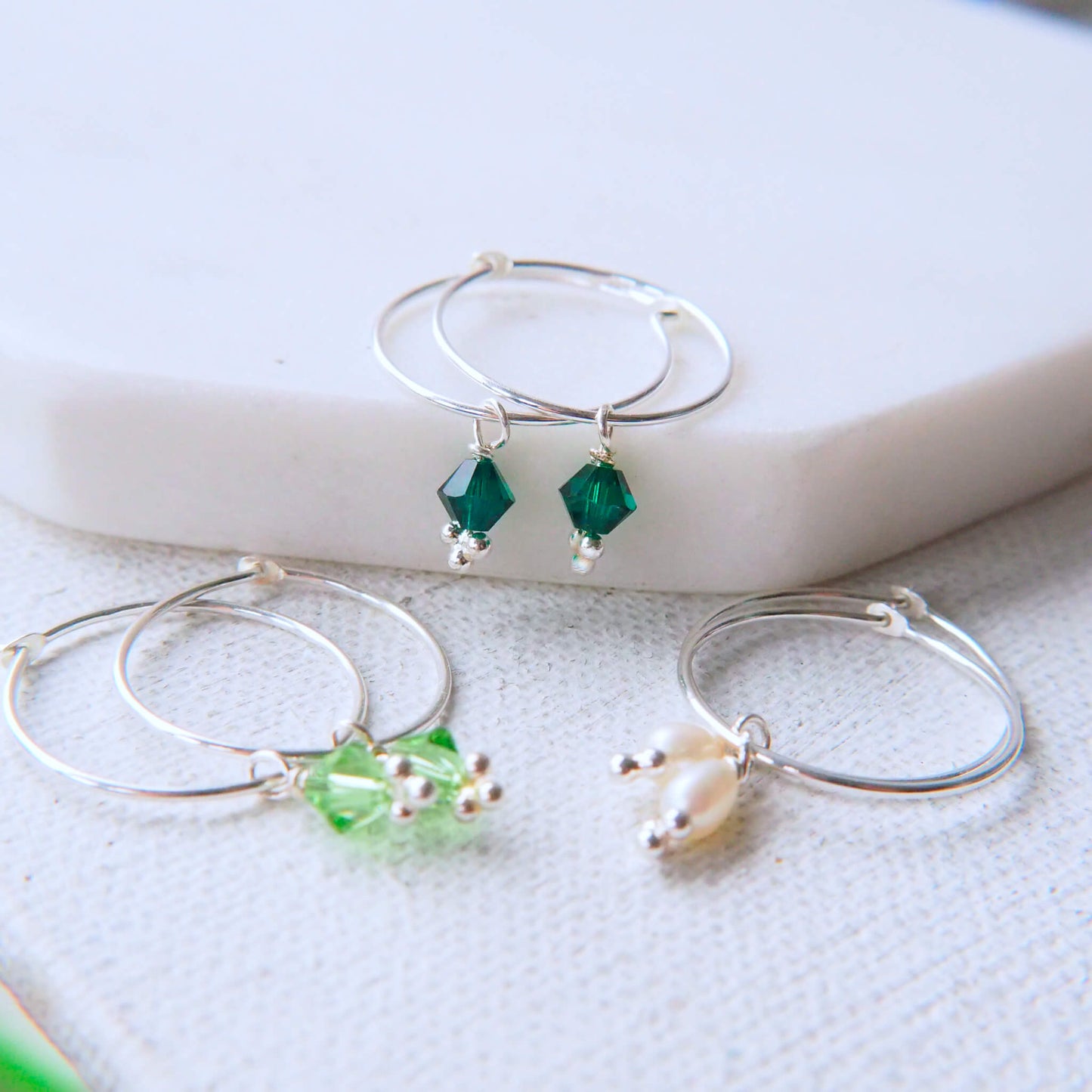 Birthstone hoops with wire hoops and a birthstone crystal pictured on a white marble background with May Emerald, August Peridot and June Pearl variations. Handmade by maram jewellery in Scotland