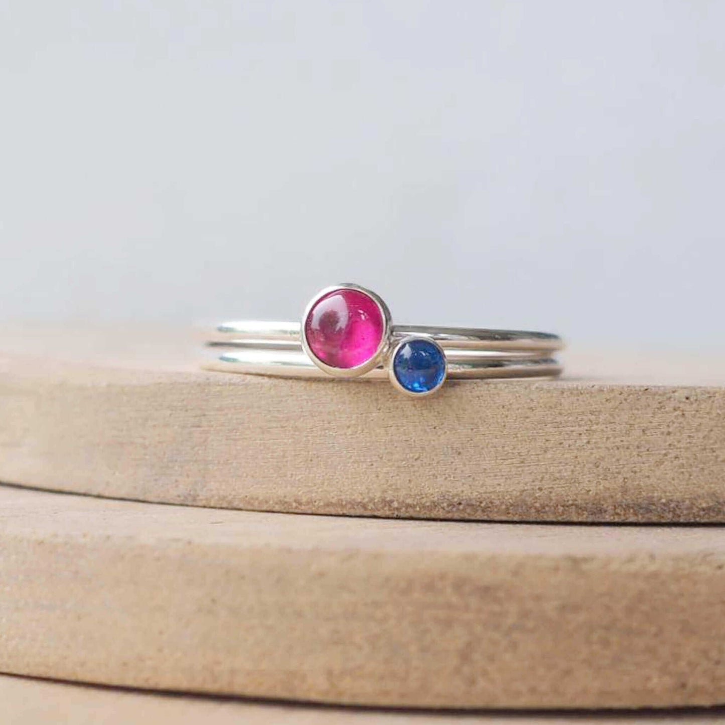 Two silver ring set with round stones. Simple round bands with a 5mm pinky red lab Ruby and a 3mm blue Lab Sapphire. Rings are pictured on wood background. Handmade by Maram Jewellery in Scotland