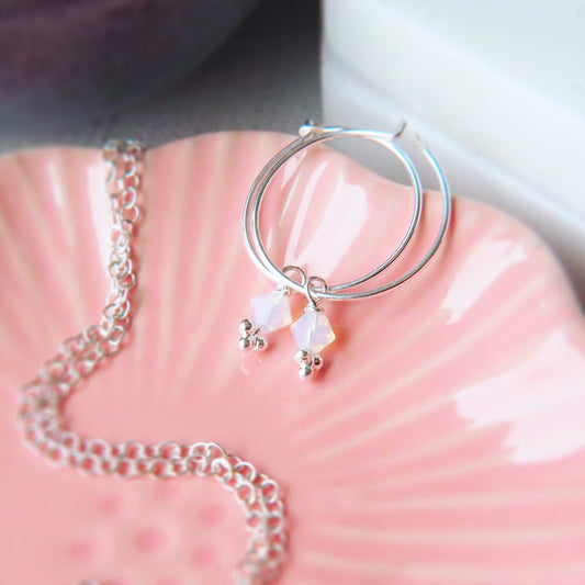 Maram Jewellery birthstone crystal earrings with opal lustre crystal. the earrings are a thin sterling silver full hoop, 2 cm diameter with a milky iridescent white crystal charm drom for October's Birthstone. handmade in Scotland