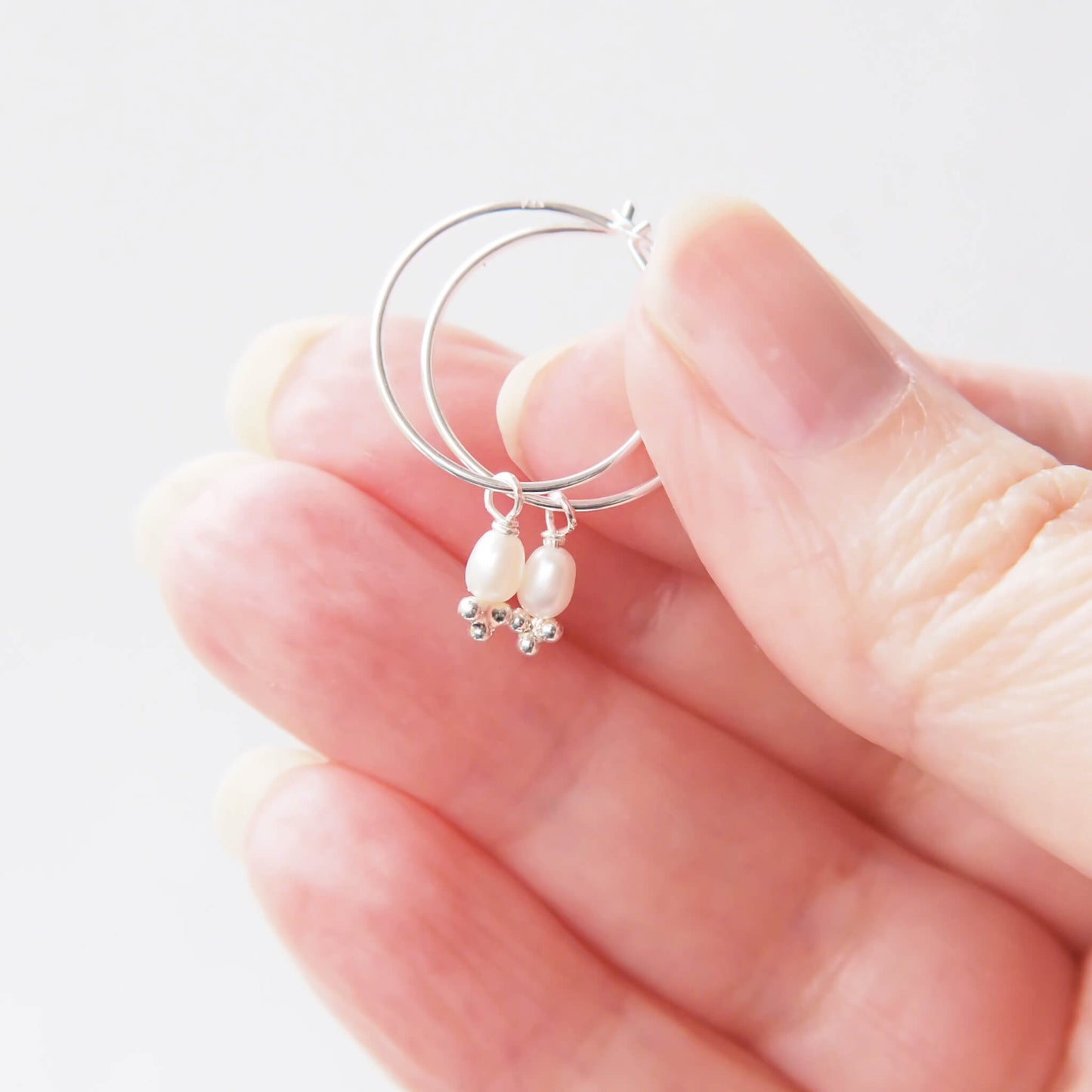 Pearl Birthstone hoop earrings held in hand to show scale. thin sterling silver wire with a freshwater pearl charm dangle. Handmade by maram jewellery in UK