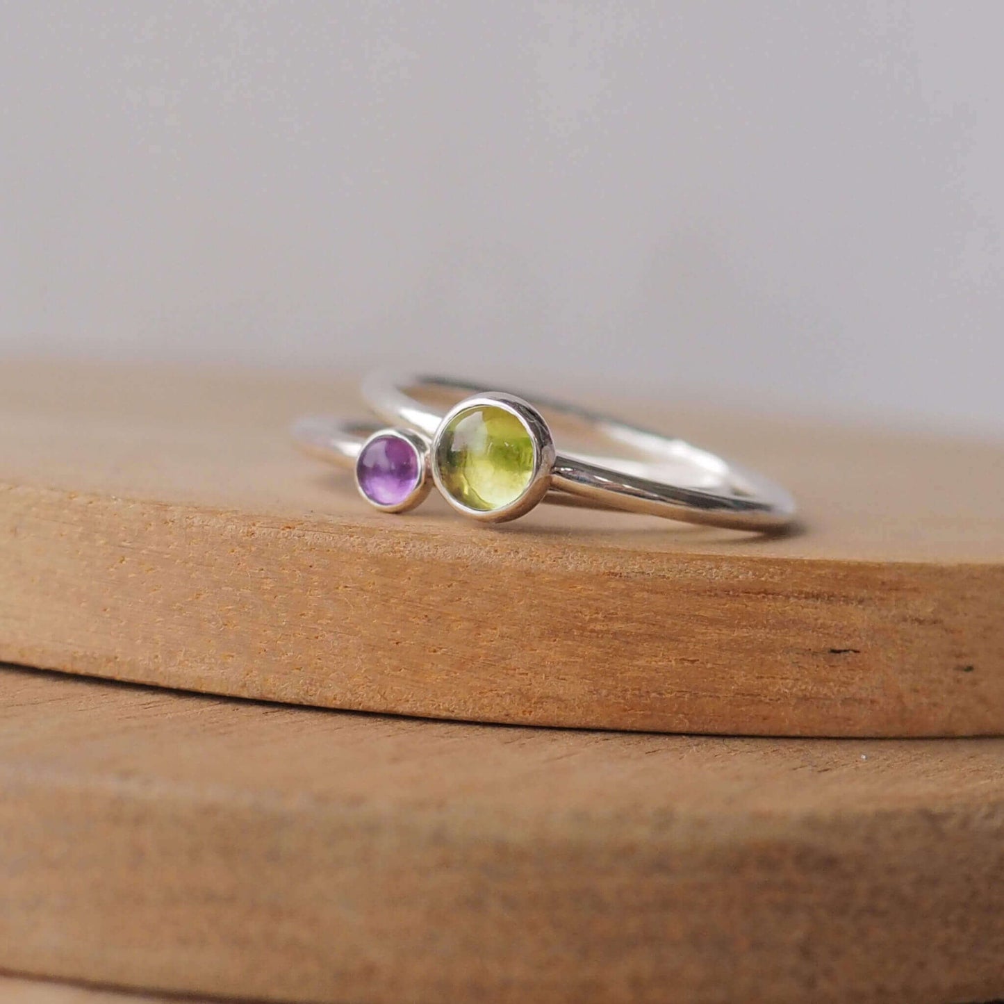Two silver ring set with round stones. Simple round bands with a 5mm green Peridot and a 3mm purple Amethyst, birthstones for August and February. Rings are pictured on wood background. Handmade by Maram Jewellery in Scotland