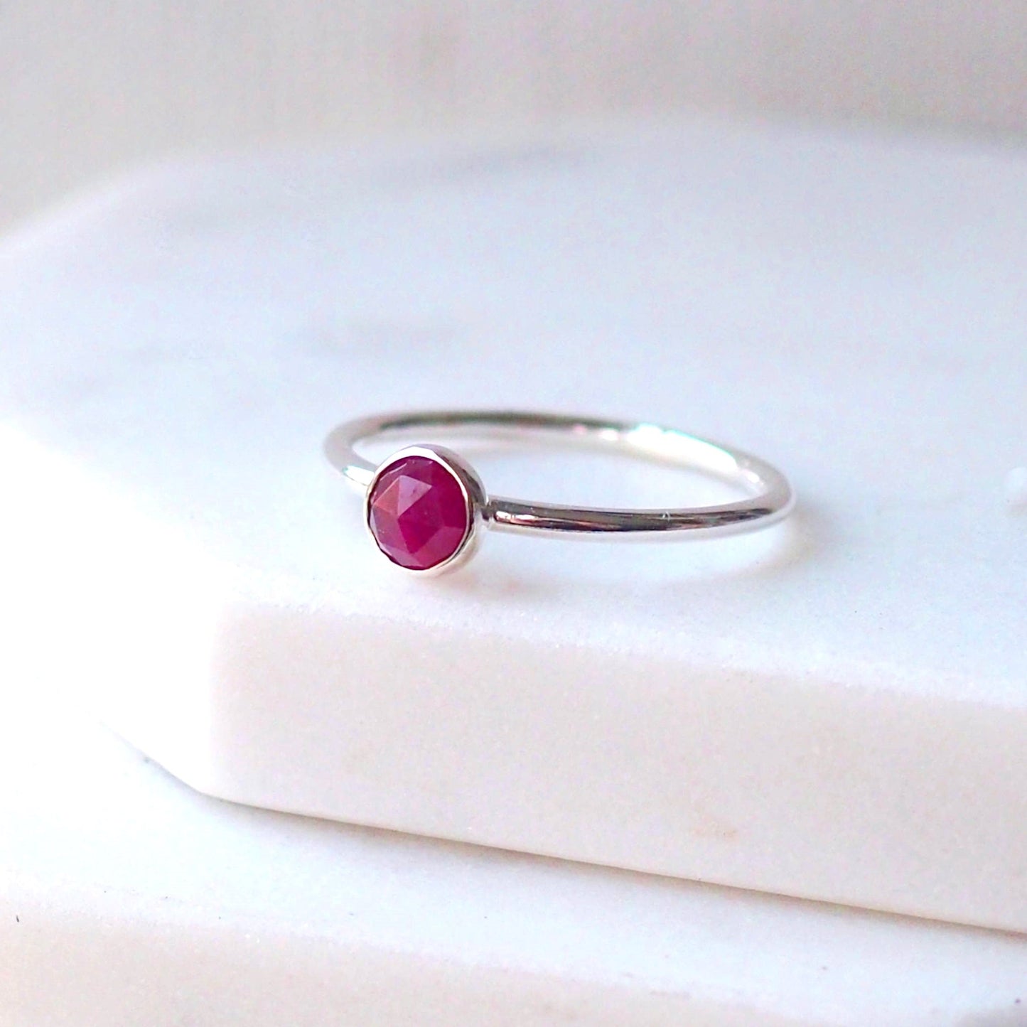 Red Ruby round gemstone ring in Sterling Silver pictured against a white marble background. The ring in minimalist in style with a round facet cut ruby in a 5mm size. Handmade by maram jewellery in Edinburgh UK