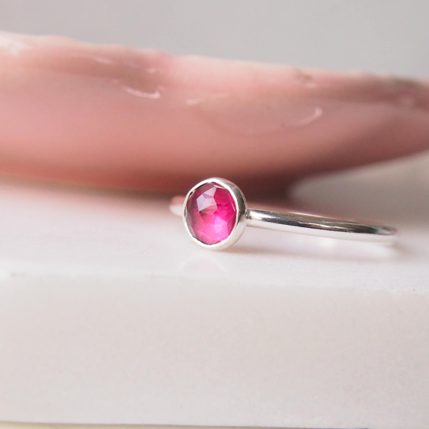 Simple raspberry rhodolite garnet solitaire ring with a 5mm round facet cabochon set onto a round halo style band. pictured on a white and pink background. Handmade by maram jewellery in Edinburgh UK