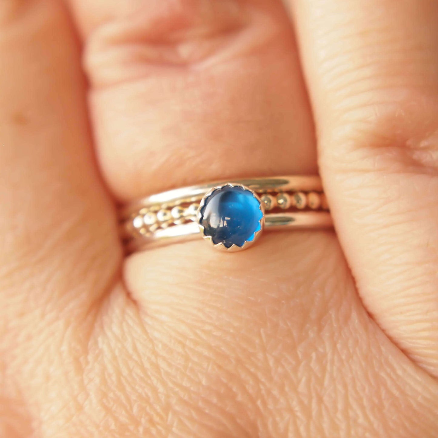 Simple styled silver and blue gemstone ring with a lab sapphire round stone on a silver band. It's shown worn on a hand with a bubble and plain silver band alongside it.The ring is made from a 5mm round cabochon in bright blue. Handmade to your choice of ring size by maram jewellery in Scotland