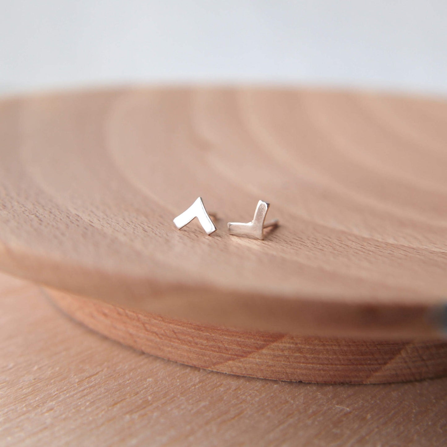 A pair of chevron tiny stud earrings in sterling silver handmade by maram jewellery. The earrings are shown on a wooden background. Chevron in shape, the studs measure 6mm in height.