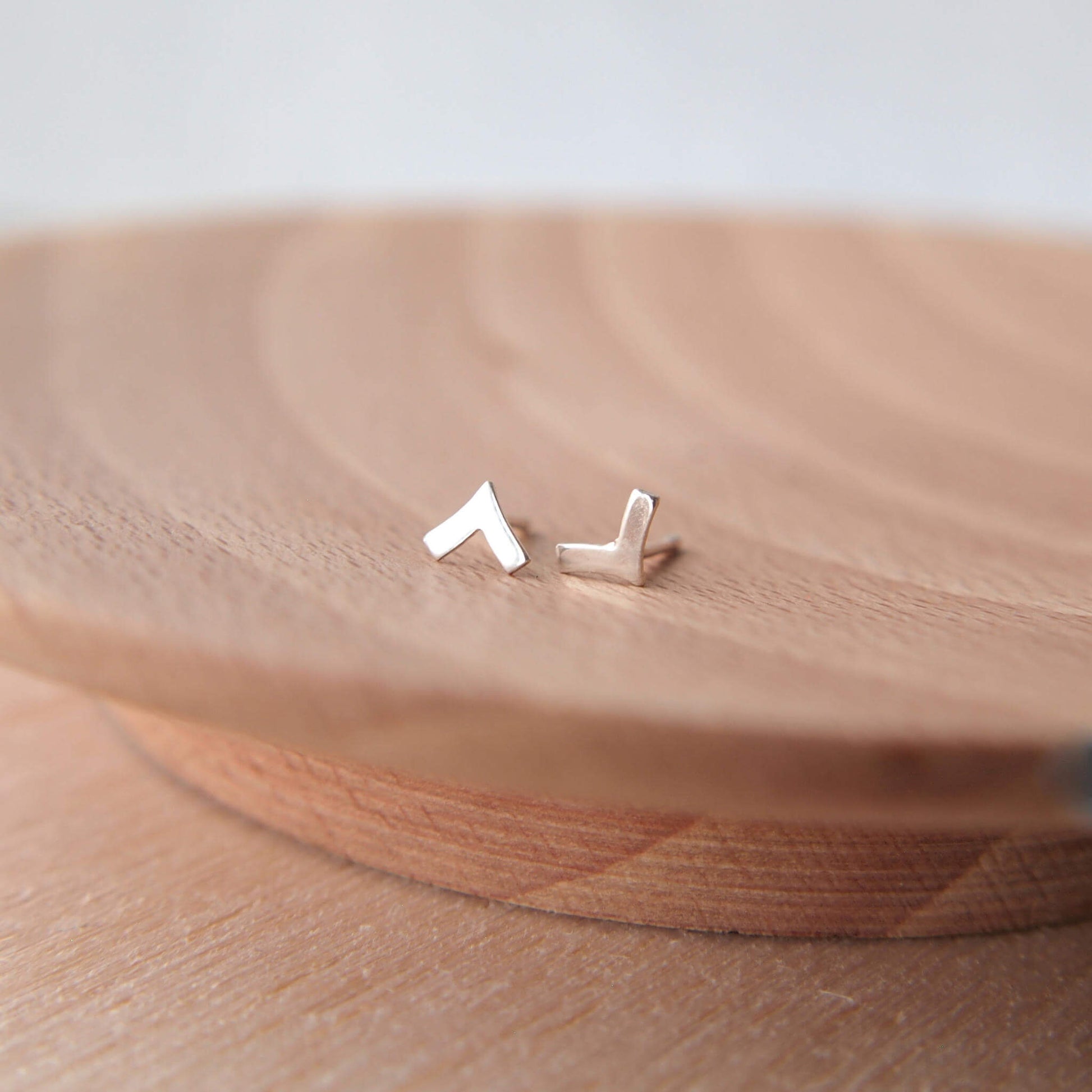 A pair of chevron tiny stud earrings in sterling silver handmade by maram jewellery. The earrings are shown on a wooden background. Chevron in shape, the studs measure 6mm in height.