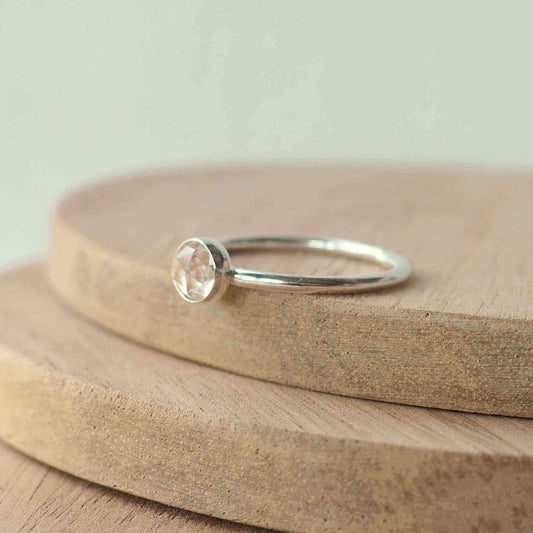 Solitaire silver ring in a modern style. Round band ring with a 5mm round white topaz with facets set very simply and minimalistic in style. Ring is on a wooden background. Handmade in Scotland by Maram Jewellery