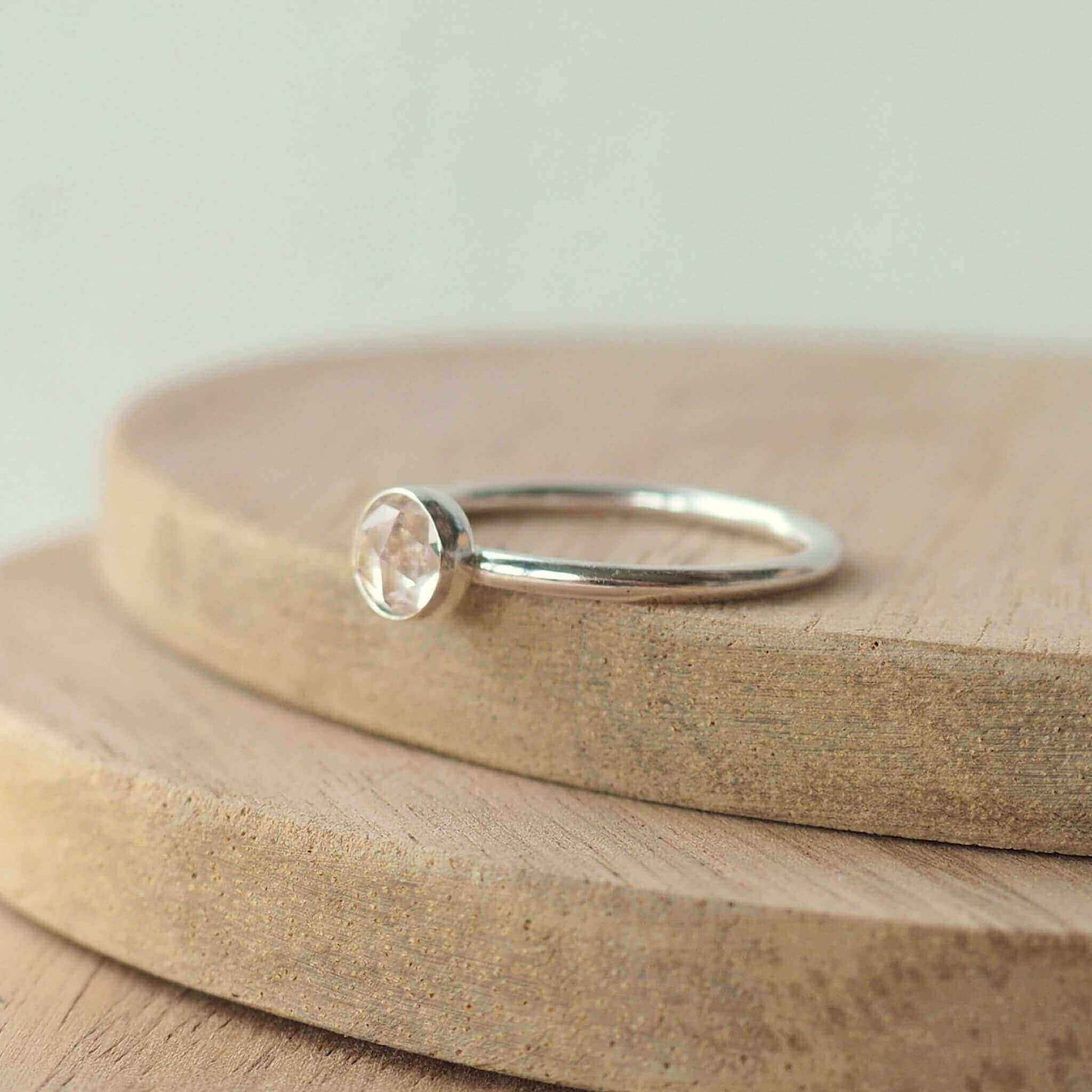 Solitaire silver ring in a modern style. Round band ring with a 5mm round white topaz with facets set very simply and minimalistic in style. Ring is on a wooden background. Handmade in Scotland by Maram Jewellery