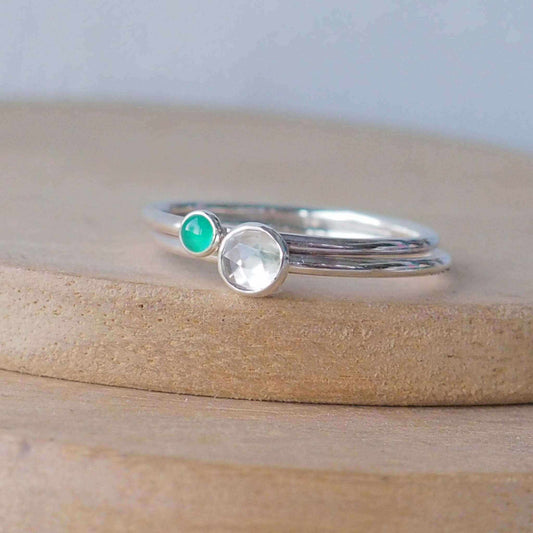 Two silver ring set with round stones. Simple round bands with a 5mm clear white topaz and a 3mm emerald green agate. Rings are pictured on wood background. Handmade in Scotland by Maram Jewellery